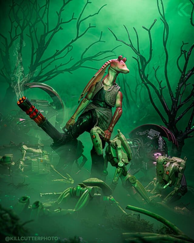 Jar Jar Binks stands triumphantly with a large futuristic gun among defeated droids in a foggy, swamp-like, green-lit scene filled with twisted branches and robotic debris.