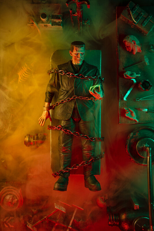 A Frankenstein’s monster figure lies strapped to a table with chains, surrounded by eerie laboratory tools and body parts, all bathed in dramatic green and red lighting with swirling mist.