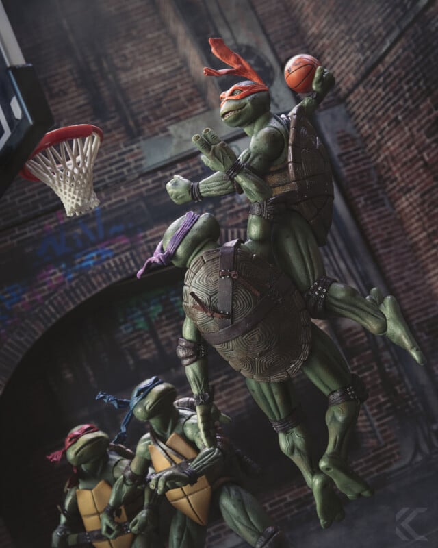 Three Teenage Mutant Ninja Turtles lift Raphael as he dunks a basketball, while Donatello and Leonardo watch in an urban alley with graffiti and a brick wall in the background.