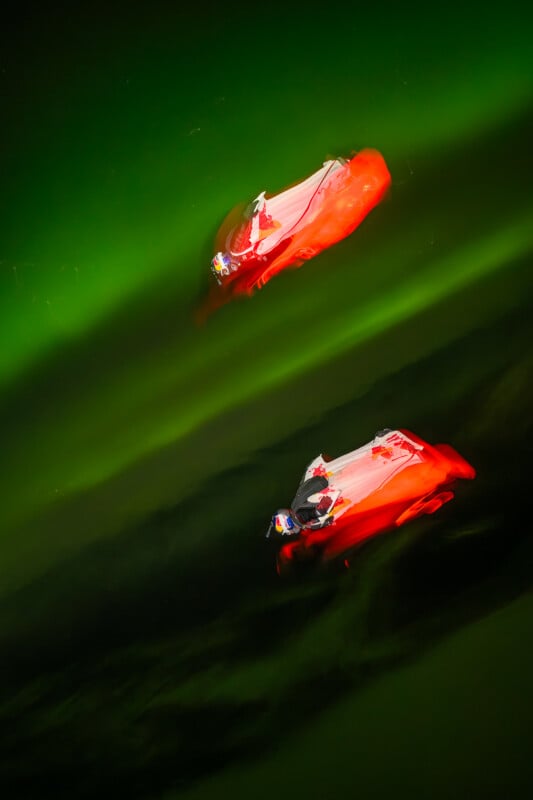 Two people in bright red kayaks paddle on dark green water at night, illuminated by vivid lighting, creating glowing reflections and a surreal, abstract effect.