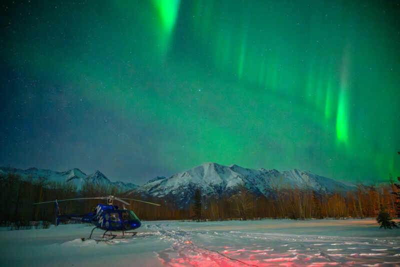 A blue helicopter sits on snowy ground beneath bright green aurora borealis lights in the night sky, with snow-covered mountains and leafless trees in the background.