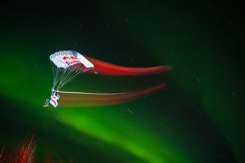 A parachutist with a Red Bull-branded parachute glides through the night sky, leaving colorful light trails, with vivid green aurora borealis and stars in the background.