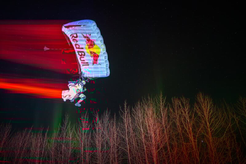 A parachutist with a Red Bull parachute descends at night above leafless trees, leaving bright red light trails behind, with a starry sky in the background.
