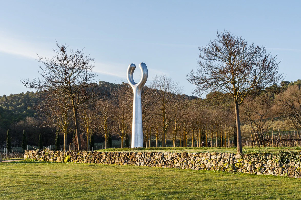Silver sculpture in a vineyard surrounded by trees and stone wall under a clear blue sky.