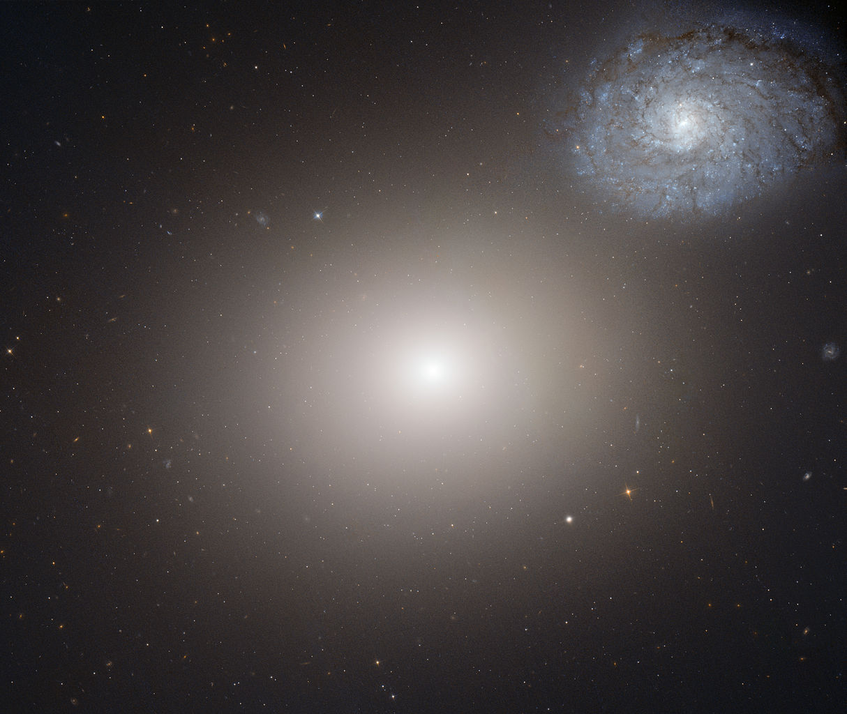 A large elliptical galaxy dominates the center, with a spiral galaxy in the upper right corner. The beauty and violence of peculiar galaxies are highlighted by the surrounding stars scattered across deep space.