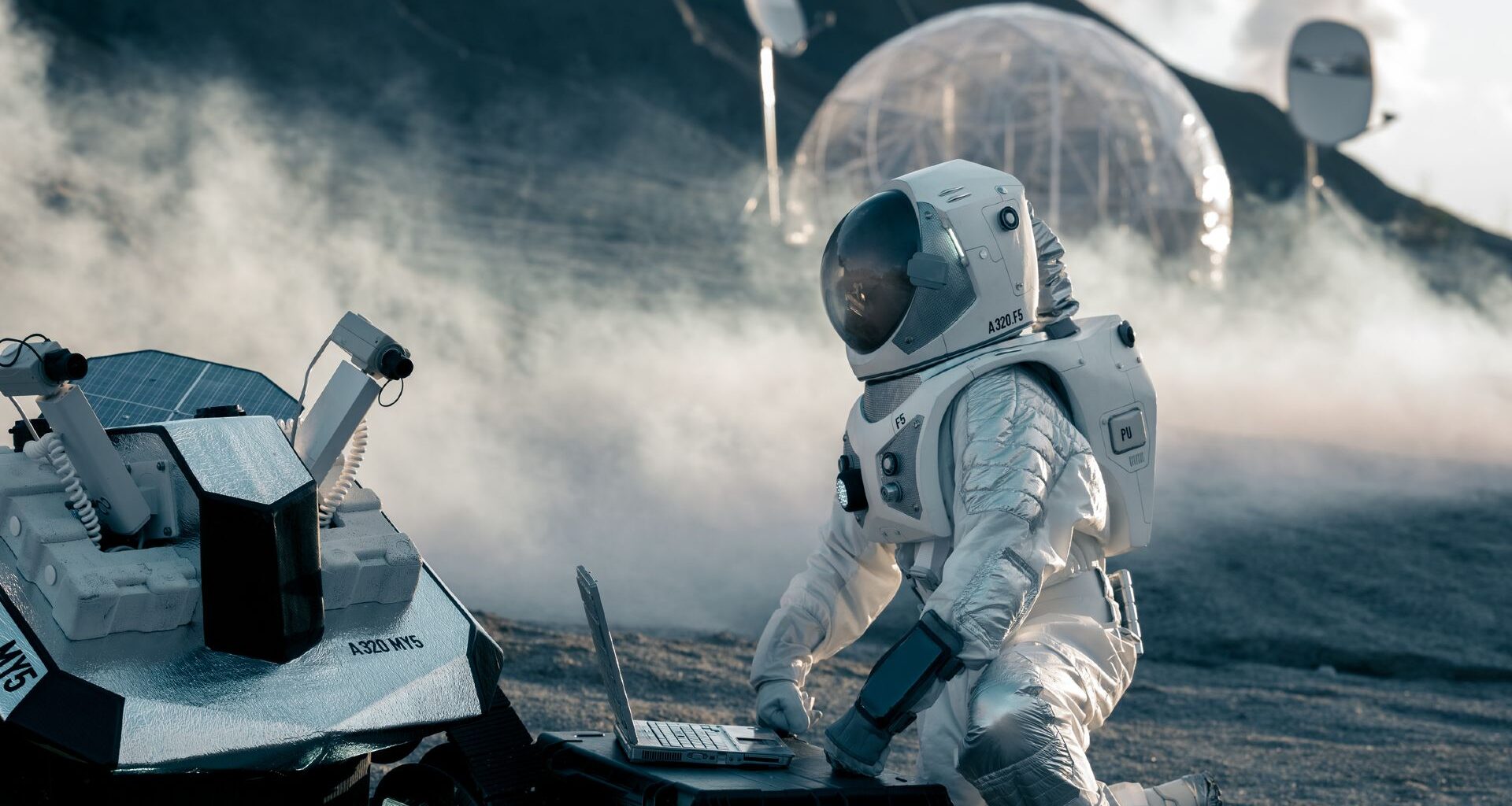 An astronaut wearing a white spacesuit kneels in front of a computer that is connected to a rover on the dusty surface of the moon with a clear circular ball behind them