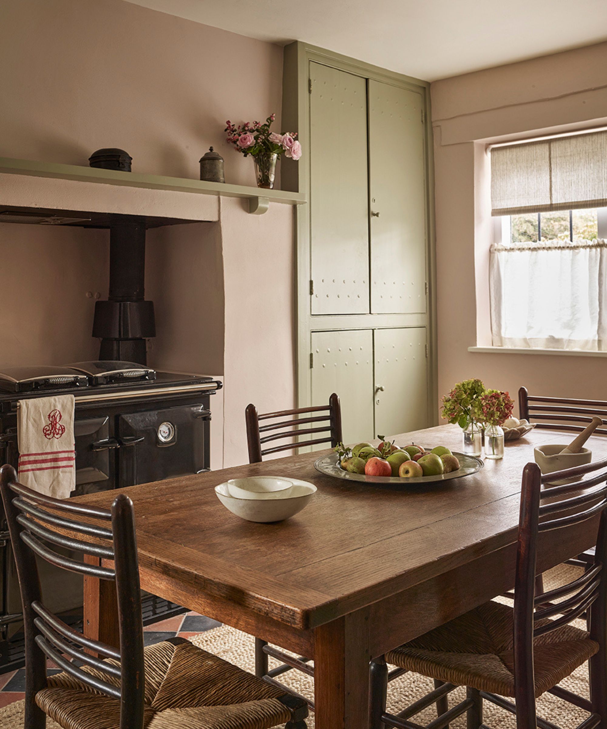 Dining room with rustic wooden table and chairs, pink walls, sage green painted cupboards, a black Aga oven, and a shelf with flowers and trinkets