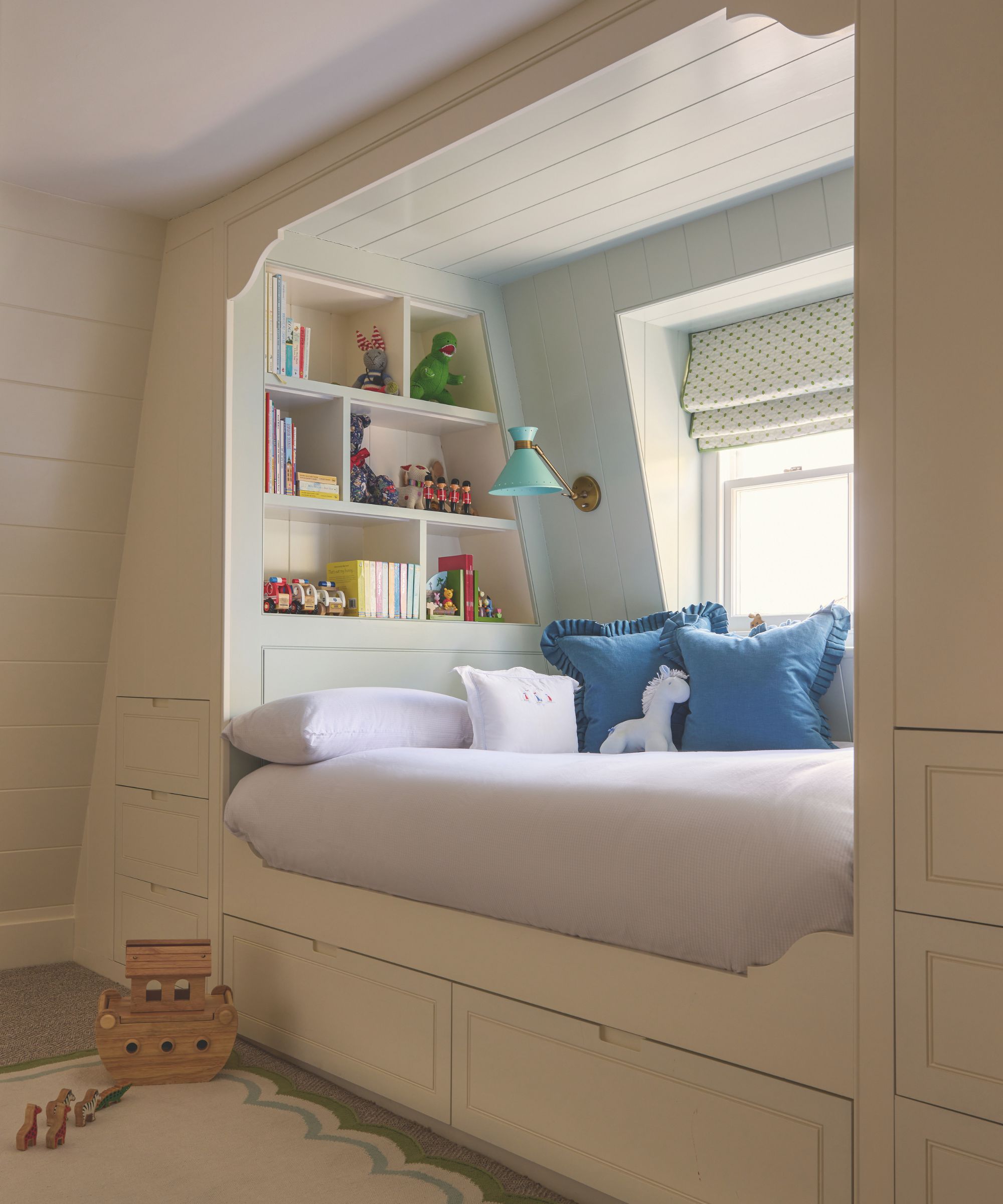 A custom-built children&rsquo;s alcove bed tucked into a cream paneled wall. The cozy nook includes built-in bookshelves filled with toys, blue ruffled cushions, a teal reading lamp, and integrated storage drawers underneath.