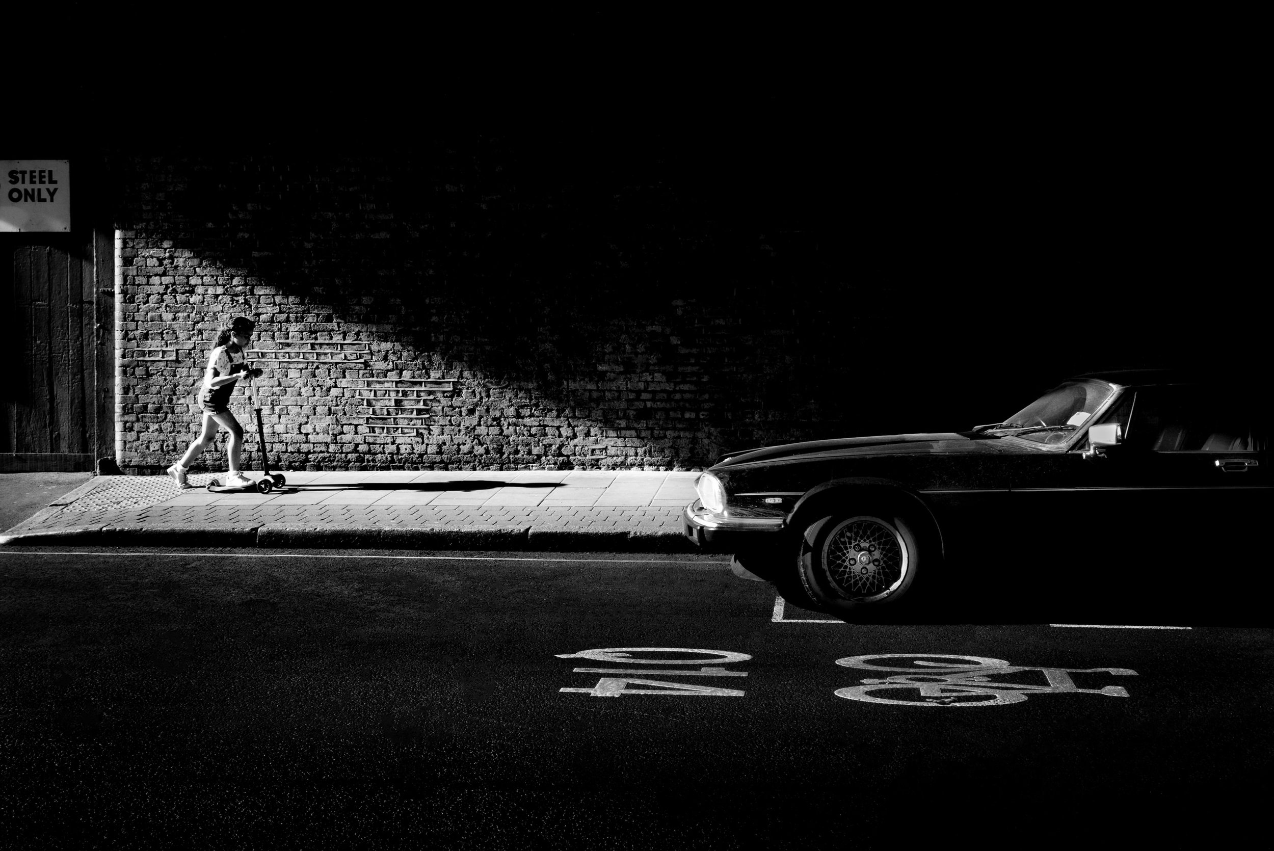 A child rides a scooter along a sidewalk in shadow, while a parked black car sits nearby, all captured in black and white