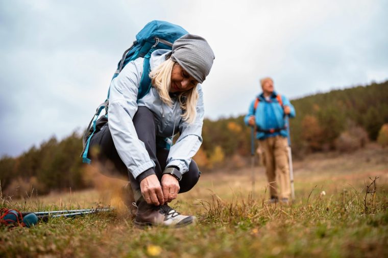 A woman is kneeling on the grass, adjusting her hiking boots while a man stands in the background, enjoying the beautiful landscape. This image captures the essence of outdoor adventure and the joy of hiking in nature.