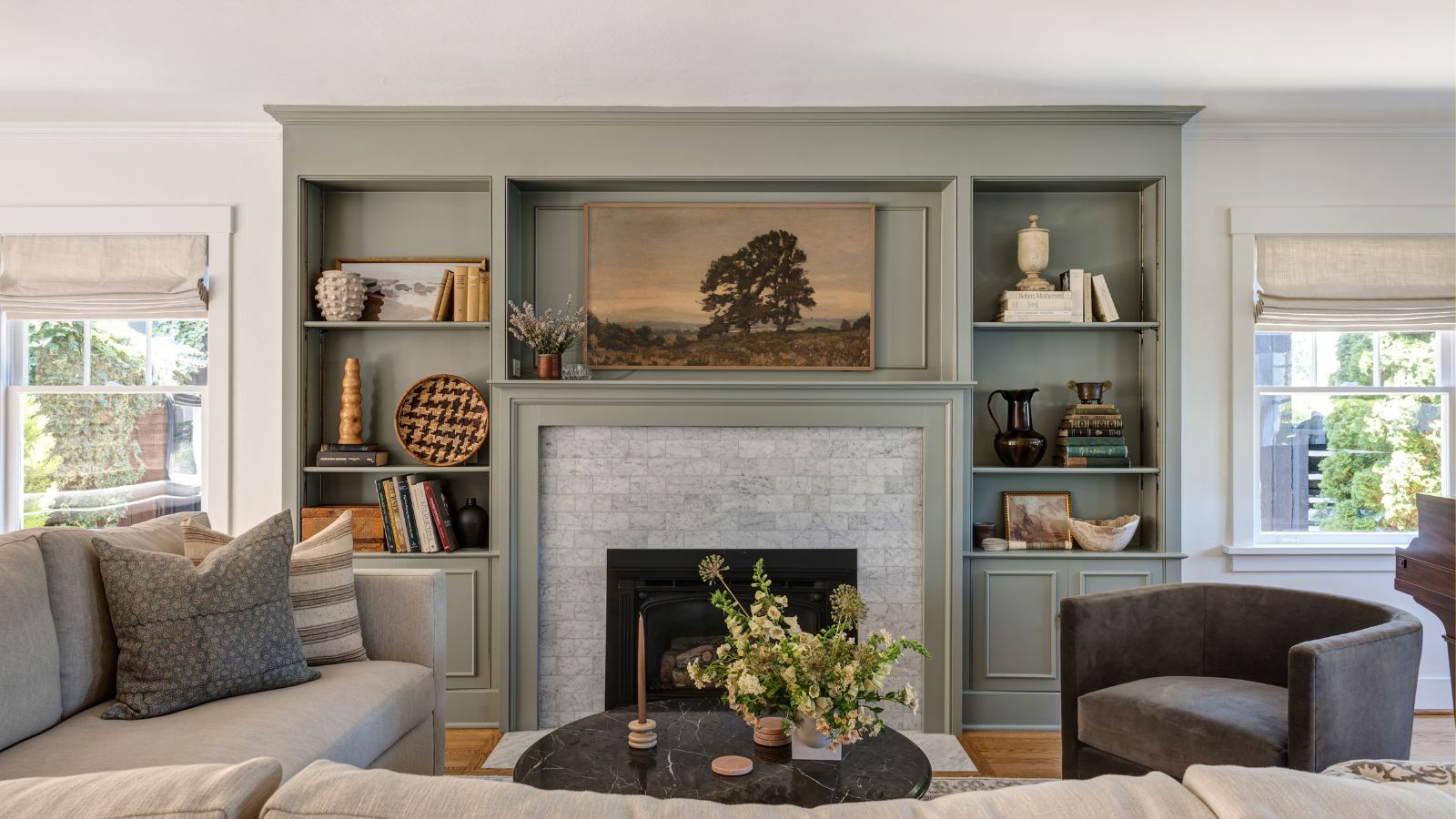 Living room with grey-green painted built-in bookshelves with decor and books, fireplace with a landscape painting above it, neutral sofa, and a dark grey velvet armchair