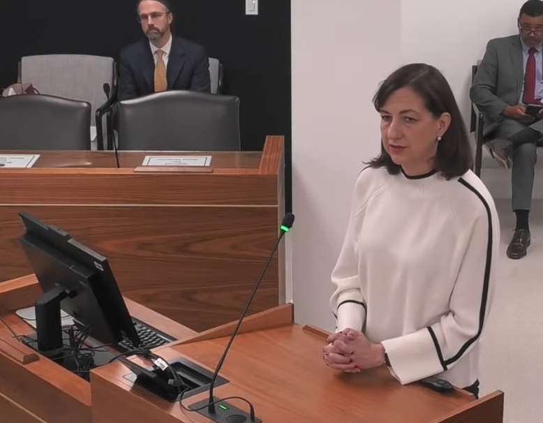 A woman stands a podium speaking at a legislative IVC committee meeting