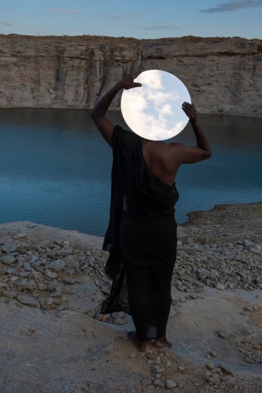 A person in a black garment stands on rocky ground by water, holding a round mirror that reflects the sky, obscuring their face and upper body. Rocky cliffs and blue water are visible in the background.