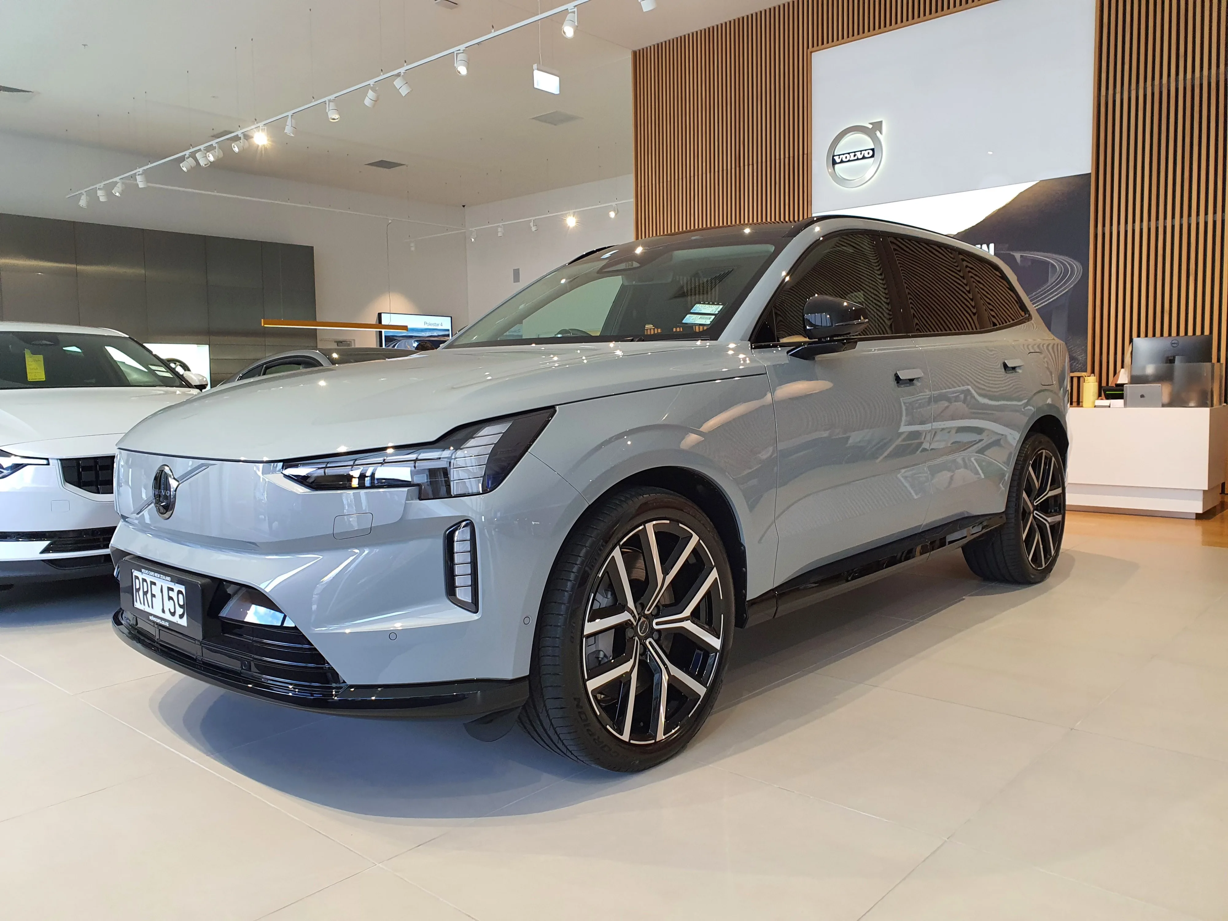 Front three quarters view of a Volvo EX90 in 'Vapor Grey' in the Archibald & Shorter Volvo North Shore dealership in Wairau Valley, Auckland, New Zealand.