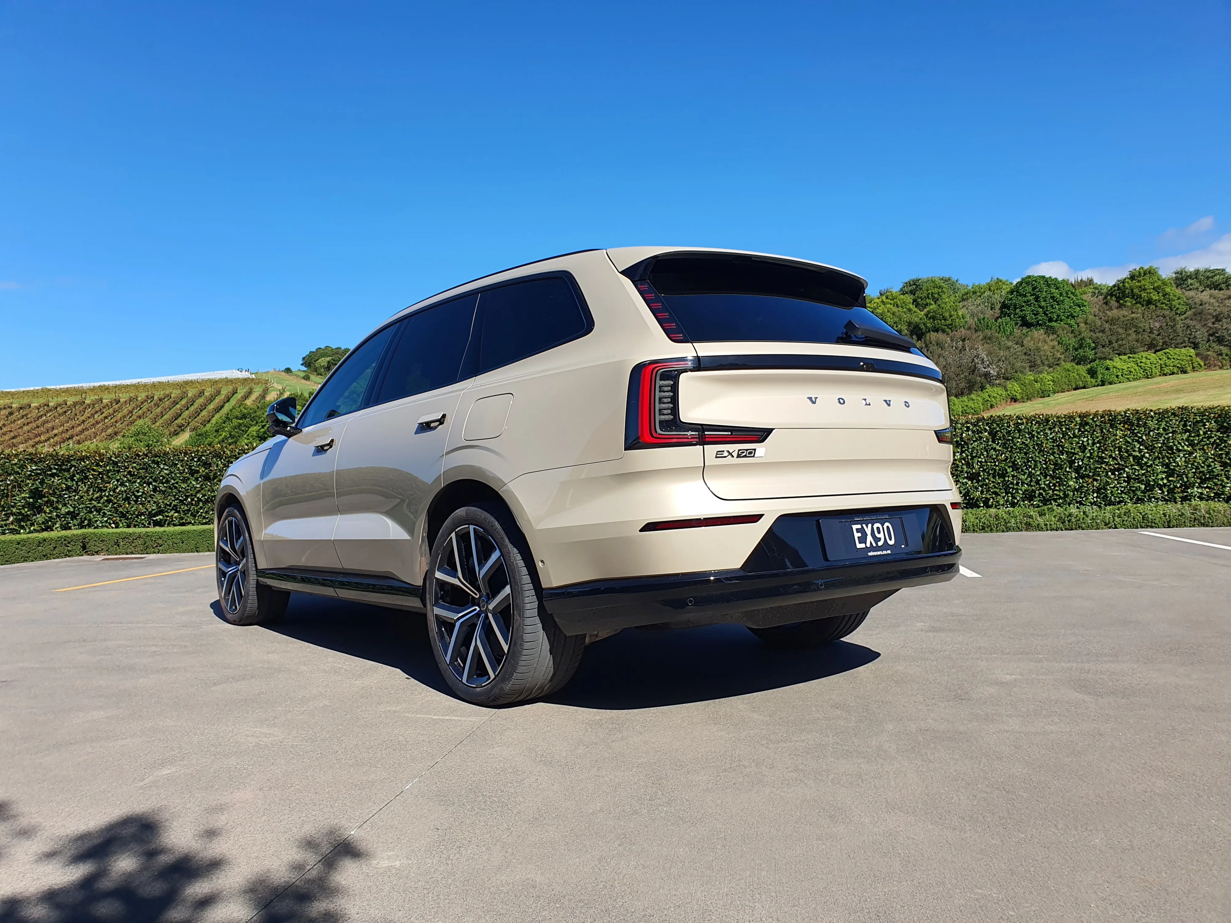 Rear three quarters view of a Volvo EX90 in 'Sand Dune' outside The Tasting Room in The Landing Winery, Purerua Peninsula, Bay of Islands, New Zealand.