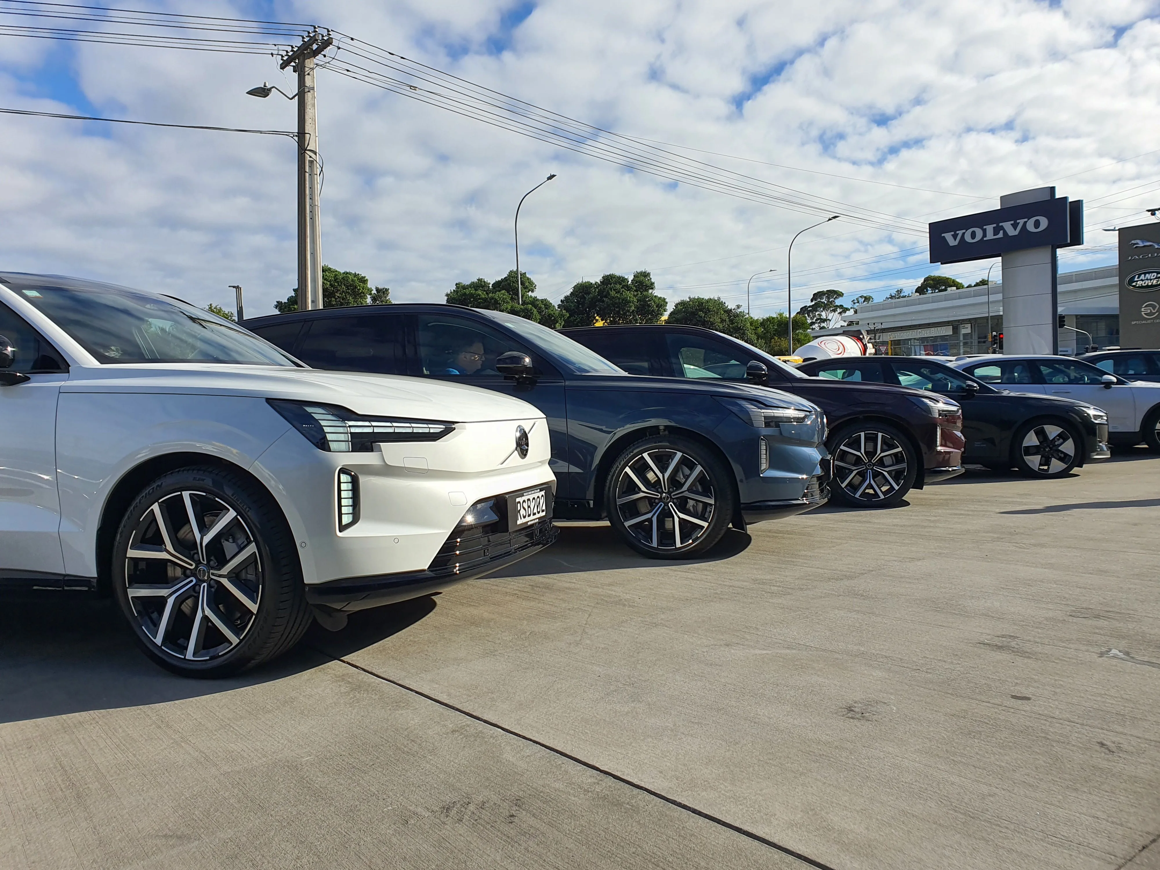 Photo of three Volvo EX90s in 'Crystal White', 'Denim Blue' and 'Mulberry Red' (left to right) in front of Archibald & Shorter Volvo North Shore dealership in Wairau Valley, Auckland, New Zealand.