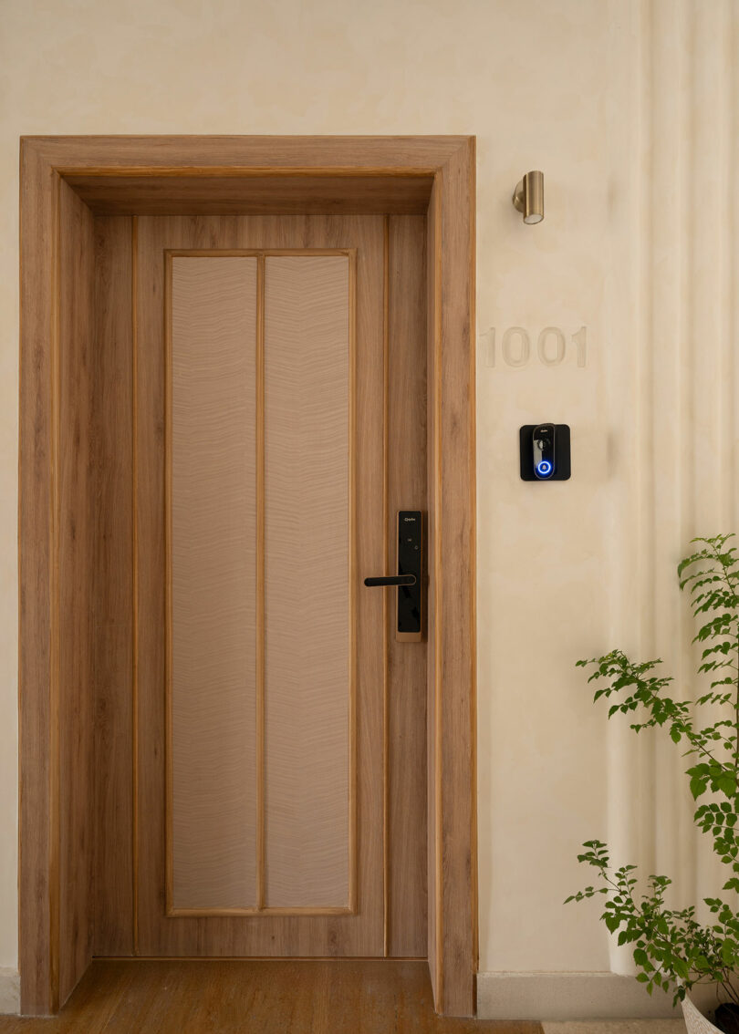 A wooden door with a black electronic lock and doorbell is set in a beige wall marked with the number 1001; a green plant is visible in the lower right corner.