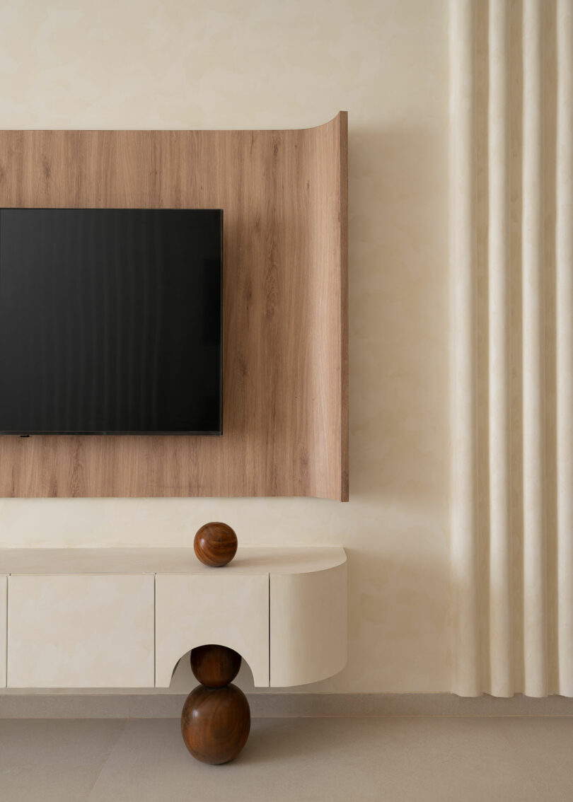 A flat-screen TV is mounted on a wooden panel above a modern white console with curved edges. Two round wooden decorative objects sit on and under the console. Cream curtains hang on the right.