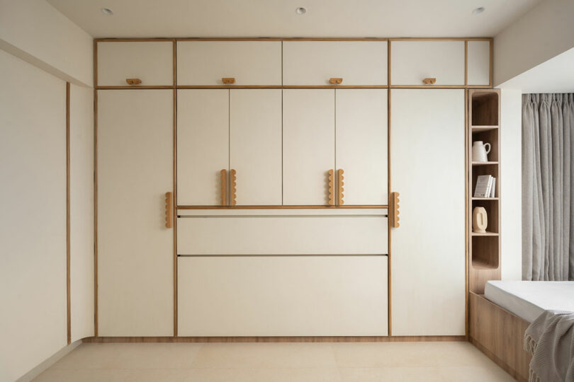 Built-in minimalist wardrobe with light wood paneling, vertical handles, upper storage cabinets, open shelves with decor, and a bed partially visible on the right.