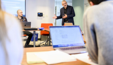 Alex Vespignani stands next to a table speaking and gesturing. Another person is seated at the table. In the foreground, another person is seated in front of a laptop.