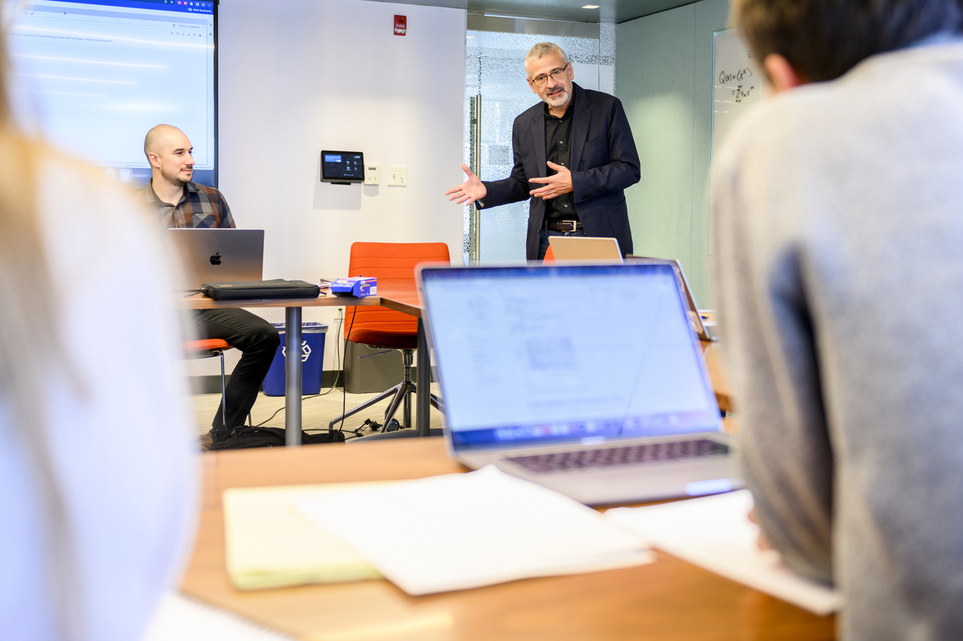 Alex Vespignani stands next to a table speaking and gesturing. Another person is seated at the table. In the foreground, another person is seated in front of a laptop.