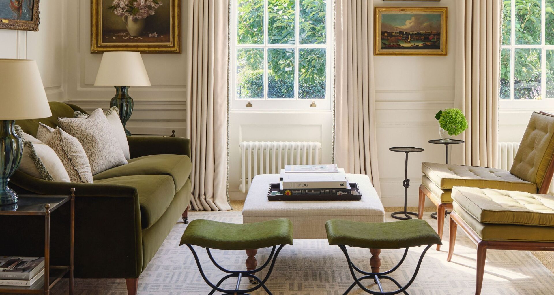 A bright, traditional living room featuring a forest green velvet sofa, olive green mid-century chairs, and twin green stools. Large sash windows are framed by cream drapes and a formal pelmet, with classical oil paintings on the walls.
