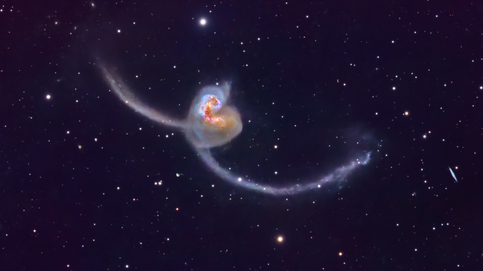 Two galaxies are pictured colliding in a dark sky dotted with stars. The orange galactic cores are visible in the center of the image and long antennae-like spiral arms can be seen stretching into space on either side.