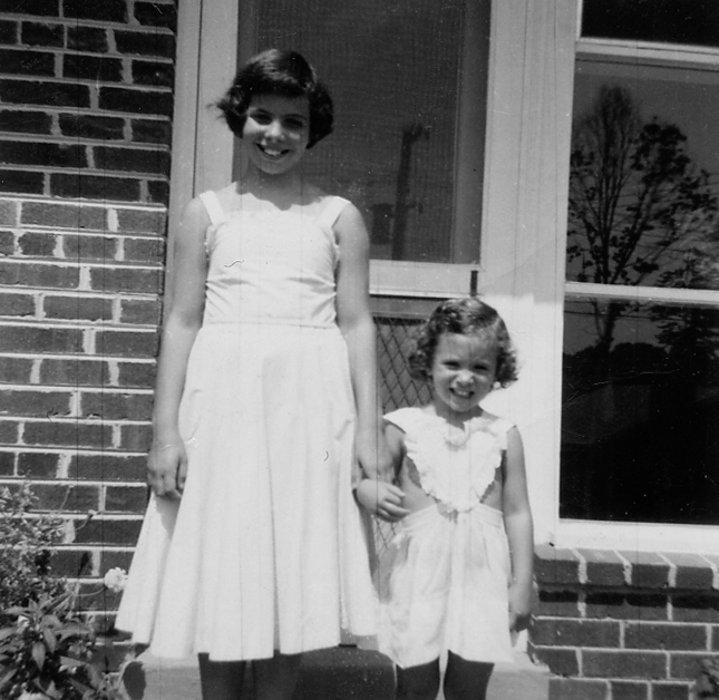 A black and white vintage-style photograph of two young girls in light-colored dresses standing together in front of a brick building.