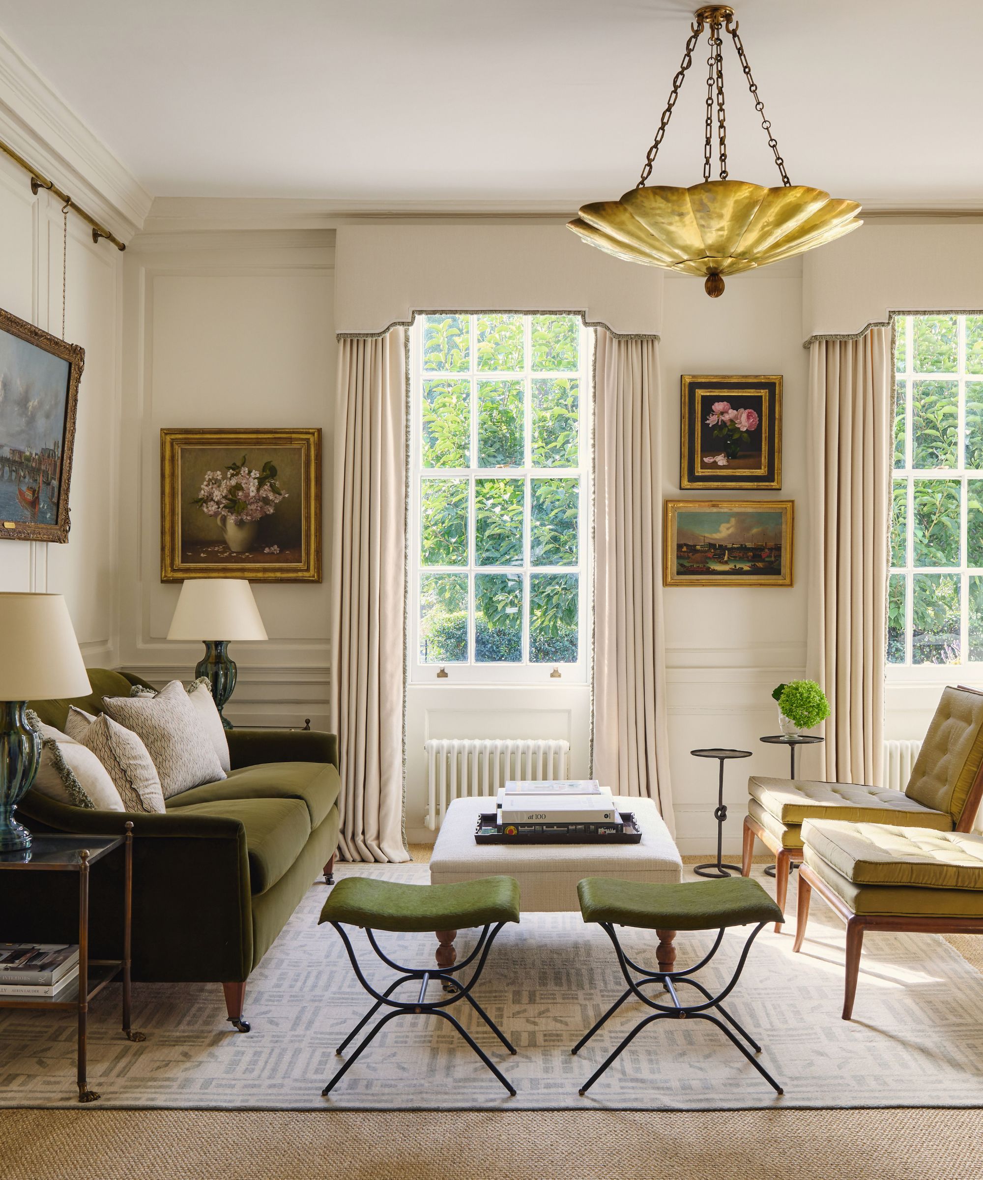 A bright, traditional living room featuring a forest green velvet sofa, olive green mid-century chairs, and twin green stools. Large sash windows are framed by cream drapes and a formal pelmet, with classical oil paintings on the walls.