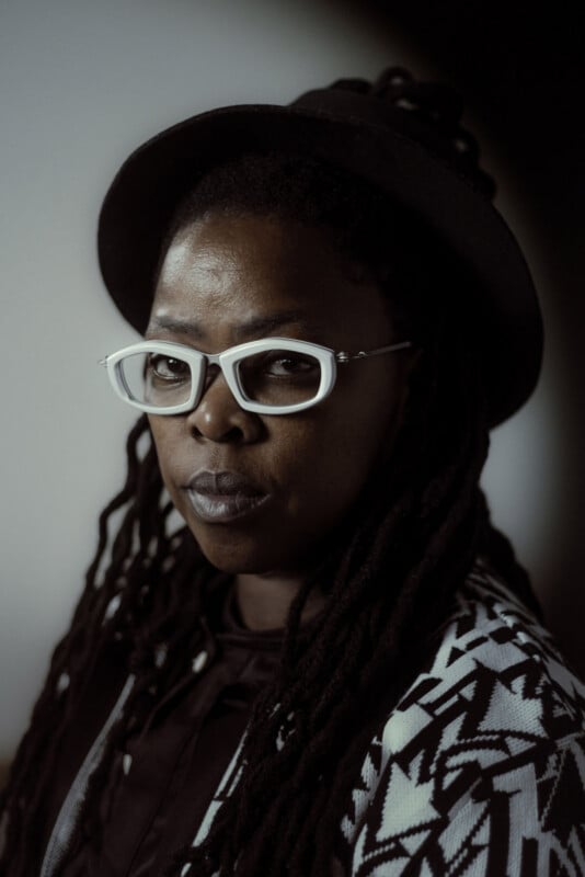 A person with long dreadlocks, wearing a black hat, white eyeglasses, and a black-and-white geometric patterned jacket, looks calmly at the camera against a neutral background.