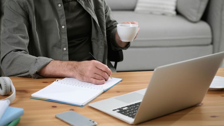 A man writes in a notebook and sips coffee while working from home.