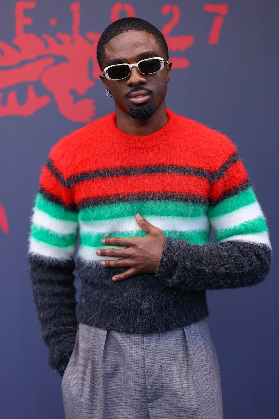 Person wearing a striped fuzzy sweater and sunglasses, posing with hand on chest, against a graphic backdrop at a celebrity event