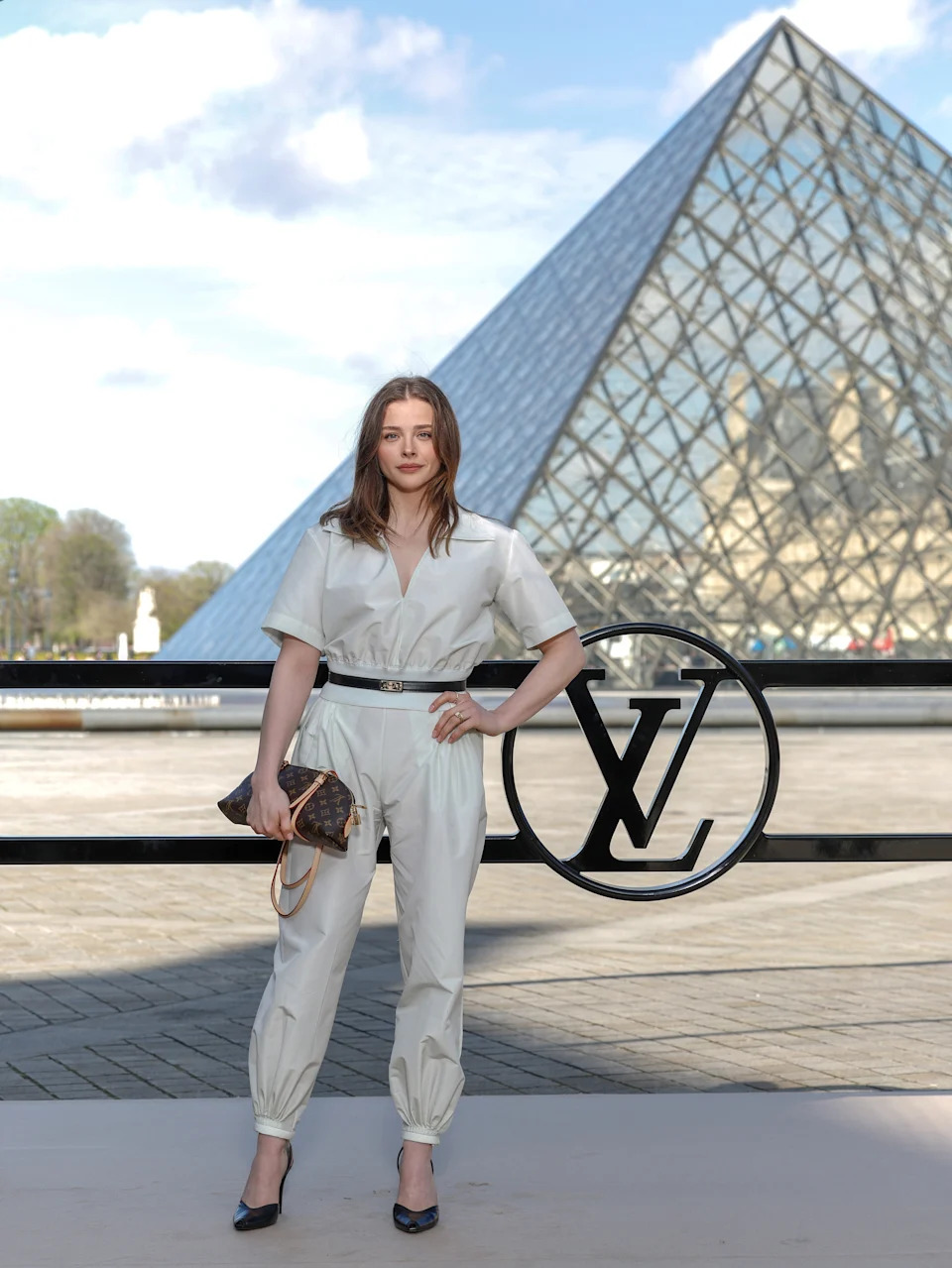Person in a stylish white jumpsuit with a black belt poses outdoors by the Louvre Pyramid, holding a designer handbag