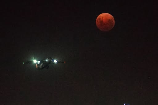 A plane flies over the Adelaide night skyline, as the moon glows red in the top right of the picture.