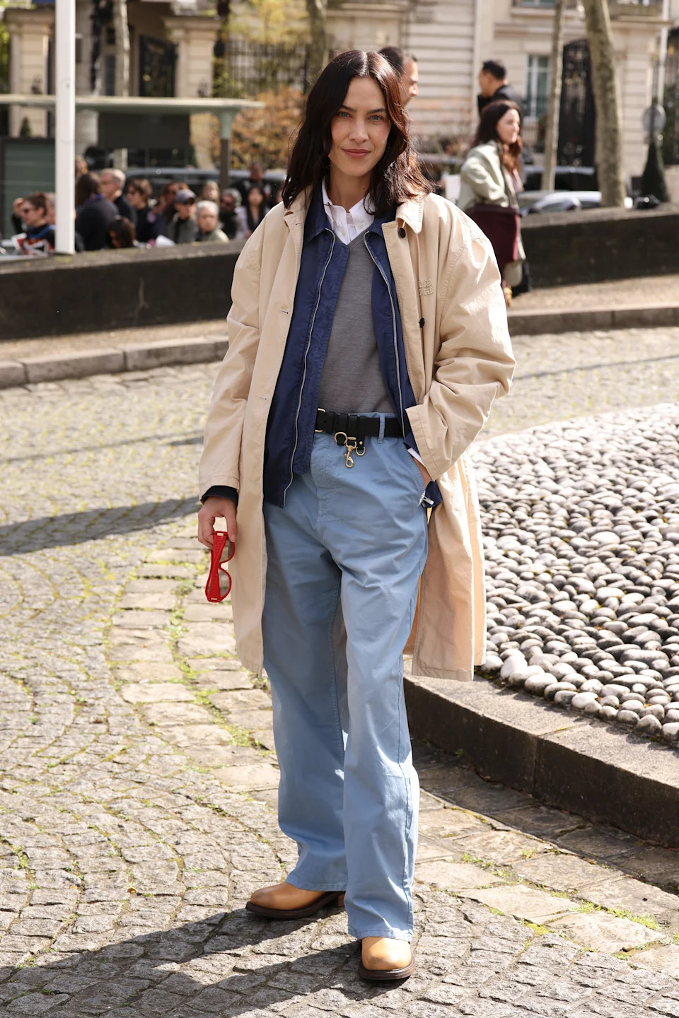 A person is wearing a long coat over a layered outfit with loose-fit pants and tan shoes, holding red sunglasses, standing outdoors
