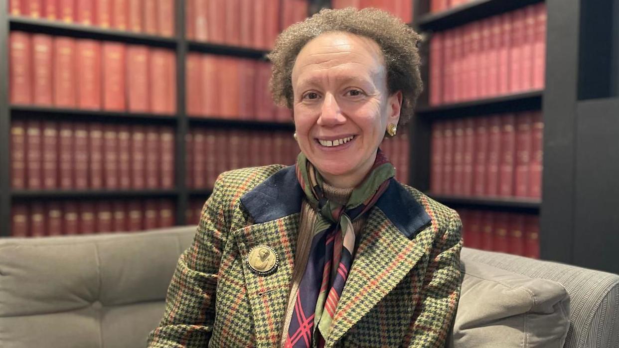 A woman in a green, navy and red checked jacket, with a similarly patterned scarf smiles at the camera. She has short, brown and curly hair and is sat on a  grey sofa in front of bookcases filled with red, leather bound books.