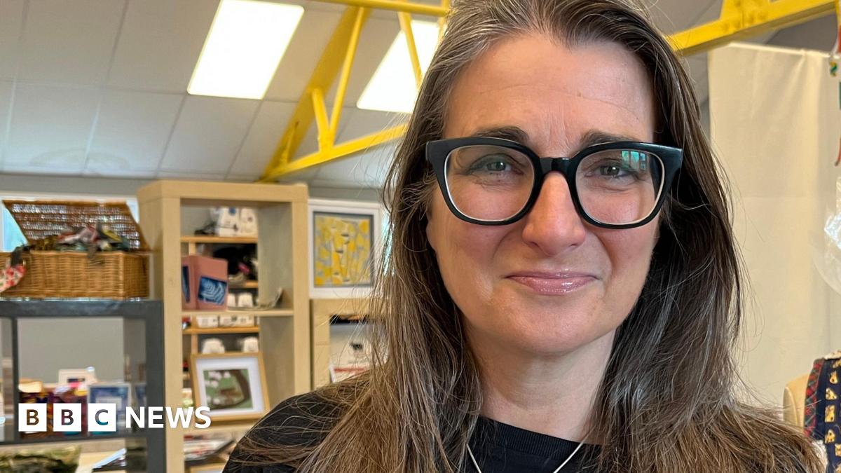 A head and shoulders image of Melissa Santiago-Val. She is wearing a black top and a necklace and glasses. She is looking directly into the camera. Behind her is some shelving with various items on it and to her left is a headless mannequin with handmade jewellery and clothing hanging on it.