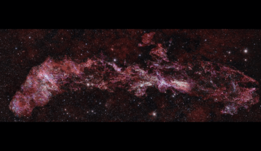 A vibrant, elongated nebula stretches across a dark, star-filled cosmic background, showcasing intricate patterns of pink, red, and purple gas and dust.