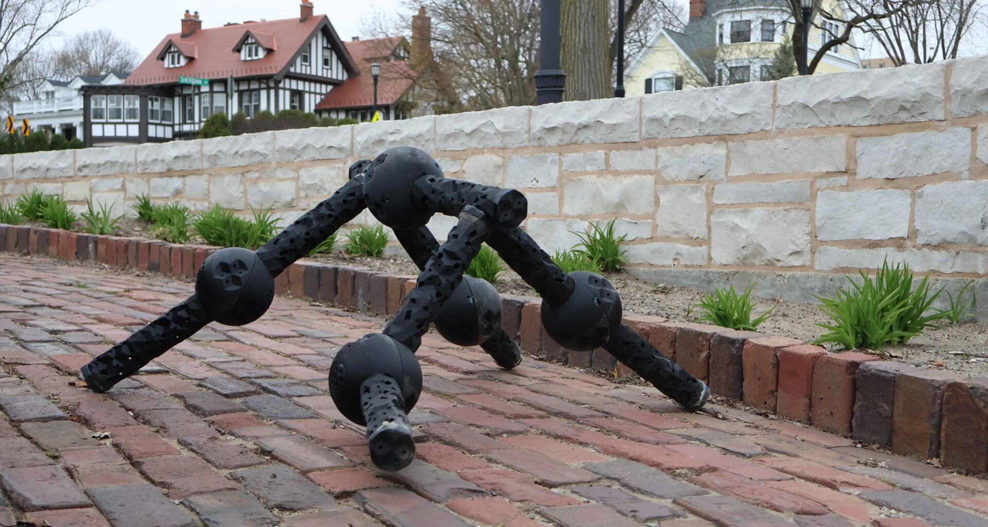 A black ball and socket robot walks on four limbs down a brick path with houses in the background