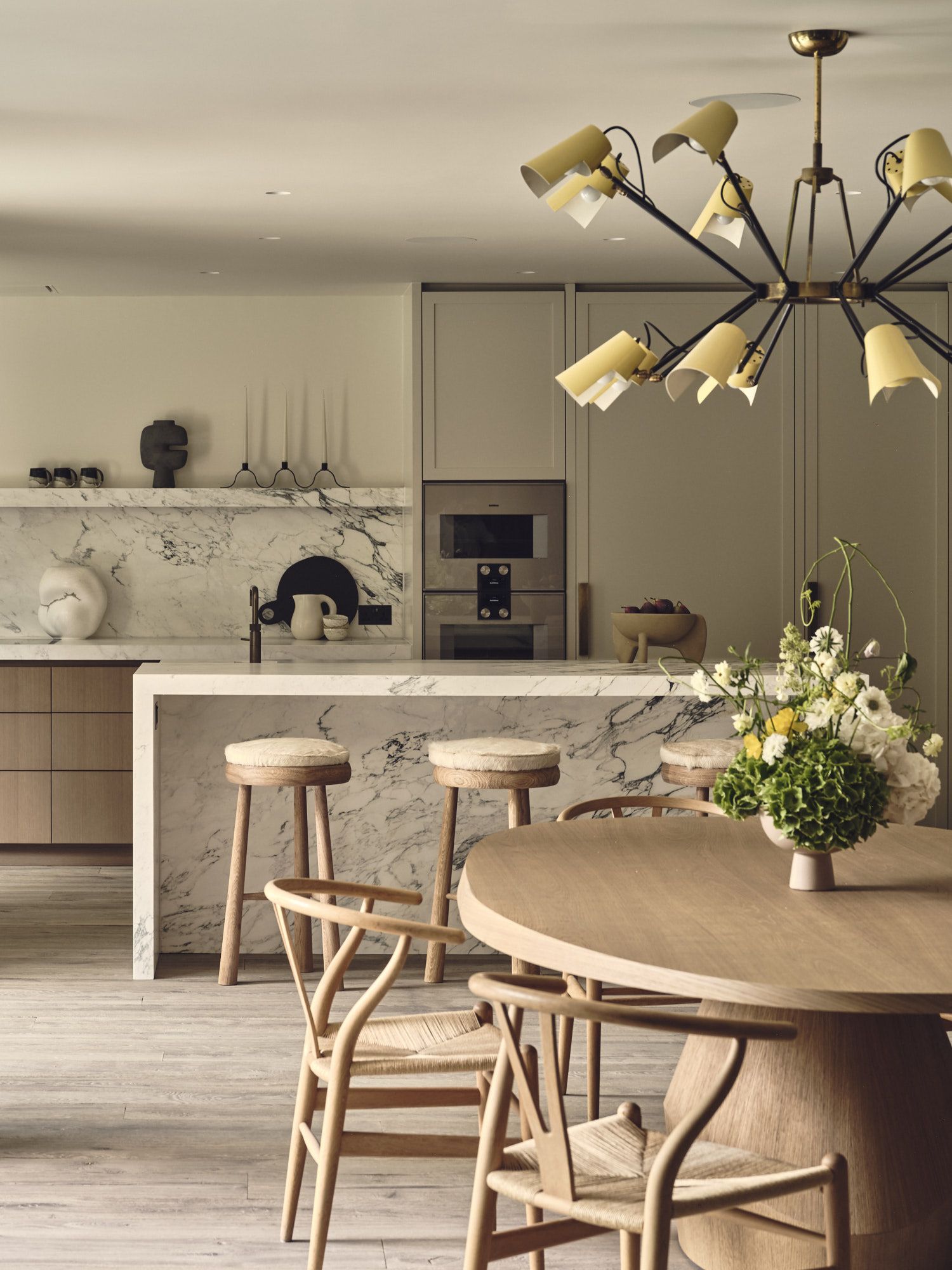 modern kitchen with stone backsplash and kitchen island and beige cabinets, sculptures on floating shelf, built-in microwave and oven, and a round timber dining table with wishbone chairs in the foreground