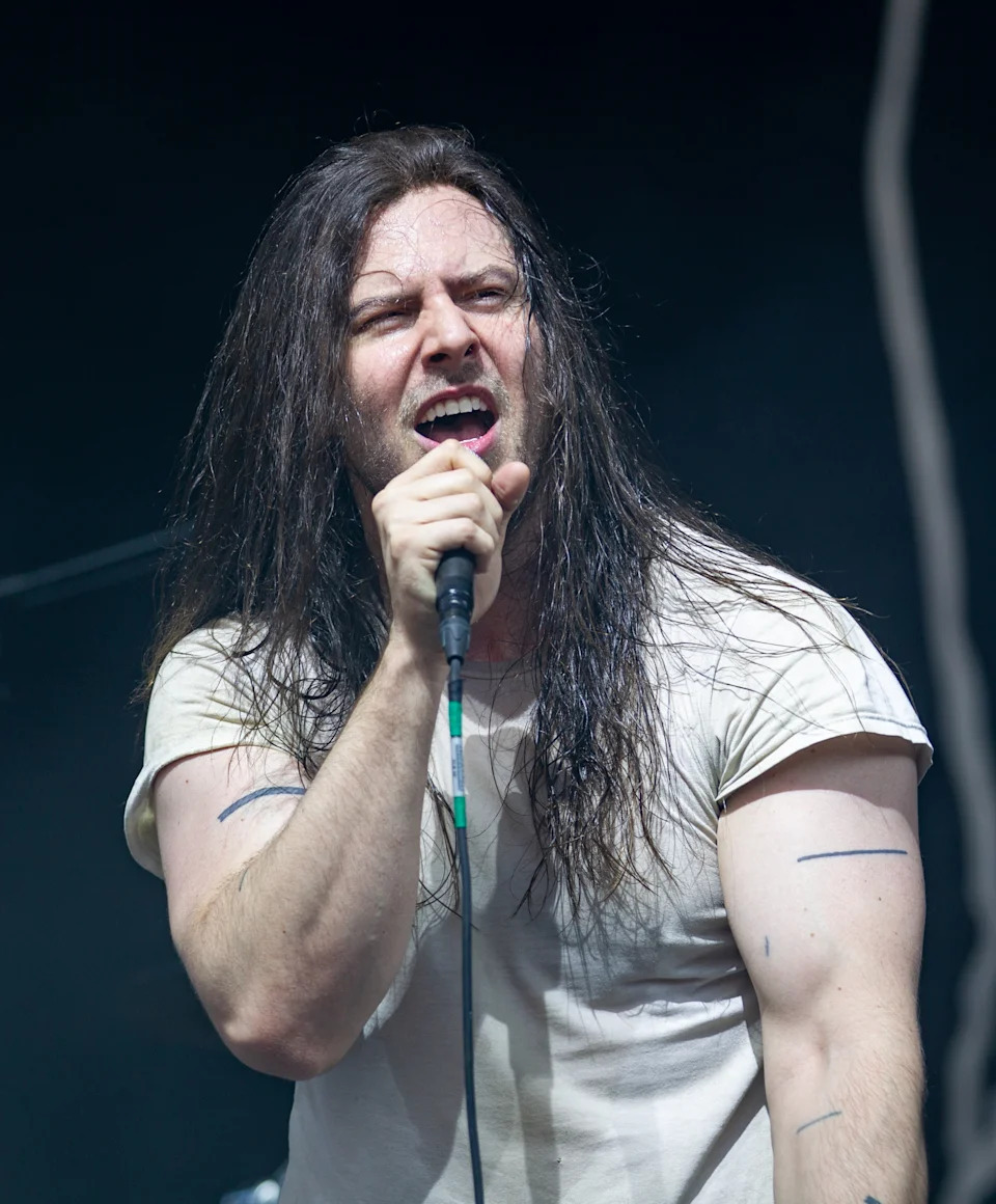 Andrew W.K. energetically sings into a microphone on stage, wearing a casual T-shirt, with long hair flowing, during a live concert