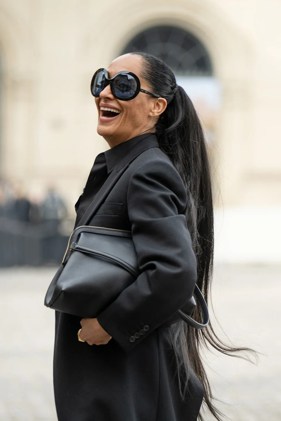 A woman with long ponytail, wearing oversized sunglasses and a sleek, black coat, smiles brightly while holding a handbag