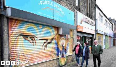 Gift Shop Purple Dragon's shutters are down. They are painted in yellow, orange and blue and who two hands reaching out to one another. Two women in jeans and dark coats are walking past and looking at the artwork. Lee Ferry is walking next to them. His hands are in the pockets of his khaki bomber jacket. He has short white hair, a beard, black circular glasses and black jeans.