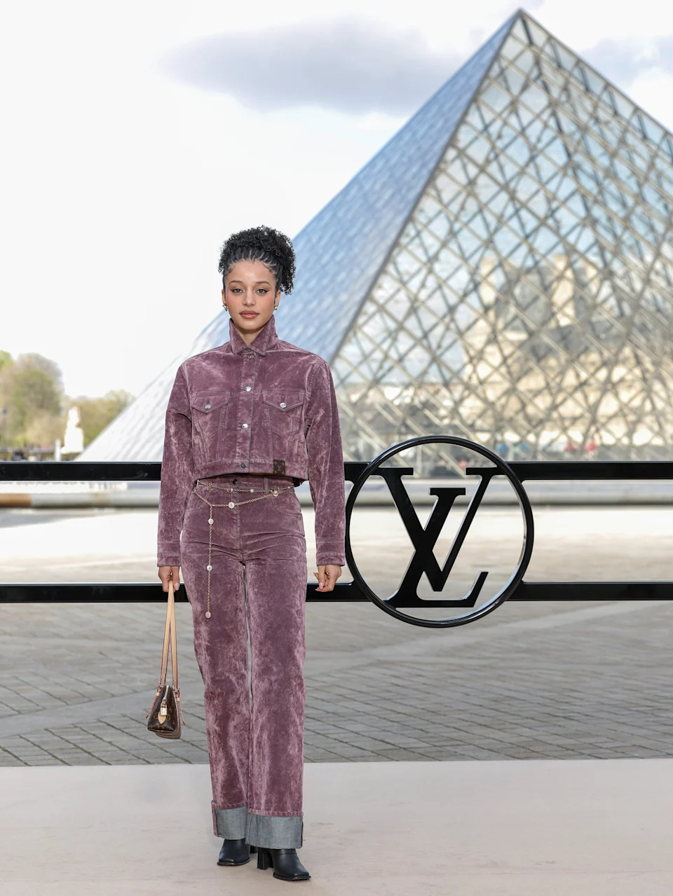 A person in a velvet ensemble stands outside the Louvre, holding a handbag near the Louis Vuitton logo