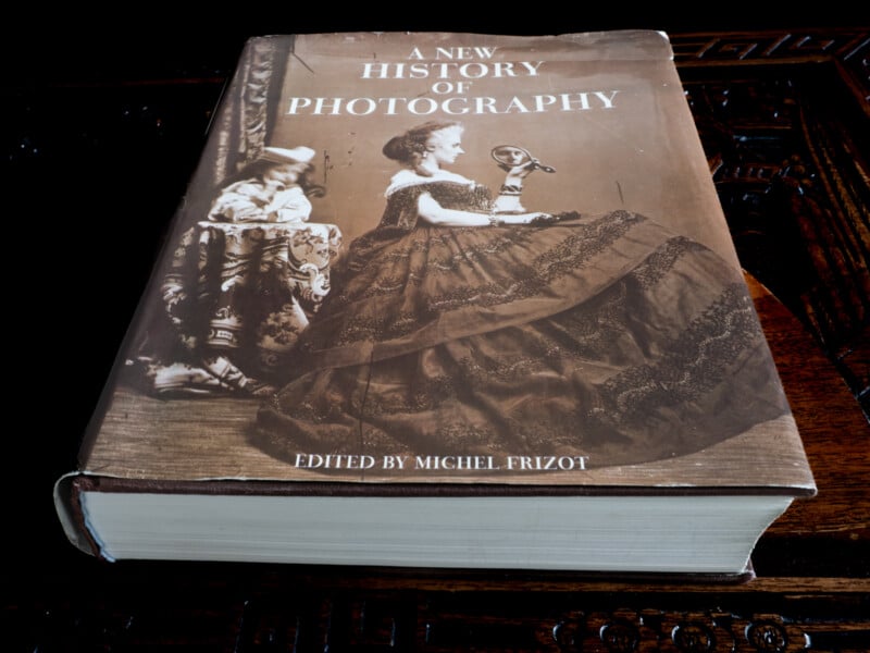 A large hardcover book titled "A New History of Photography," edited by Michel Frizot, lies on a wooden table. The cover features a sepia-toned vintage photograph of a woman in a dress holding a camera.