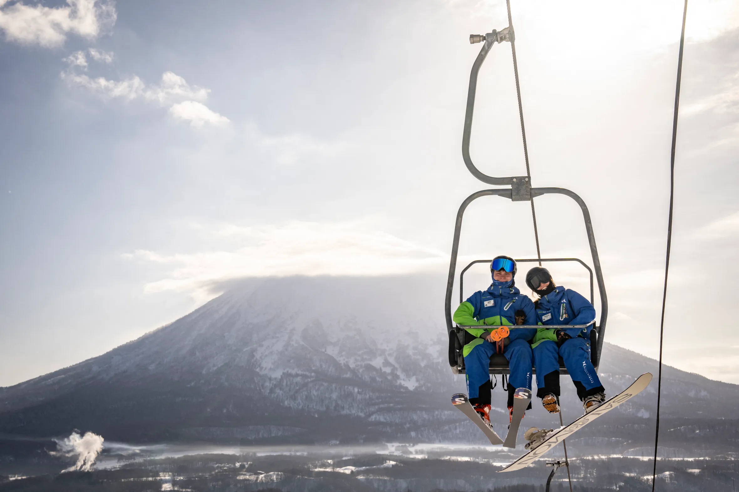 Two skiers on a ski lift with a snow-covered mountain in the background.