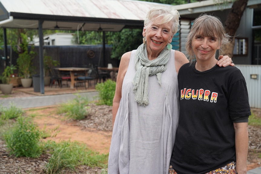 A woman rests her arm around the shoulders of another woman. They are smiling for the camera.
