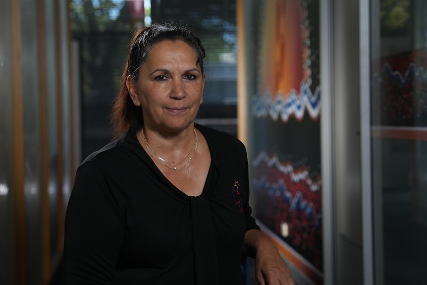 A woman wearing a black top in front of an Indigenous artwork.