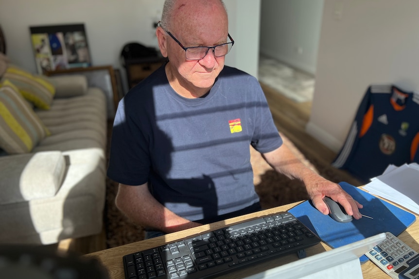 An older man in glasses sits at a computer in a home office.