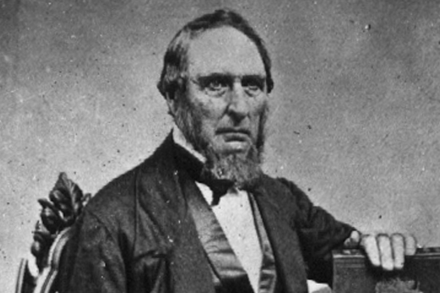 An old black and white photograph of a man with a chin-strap beard and three-piece suit sitting in a dining chair.